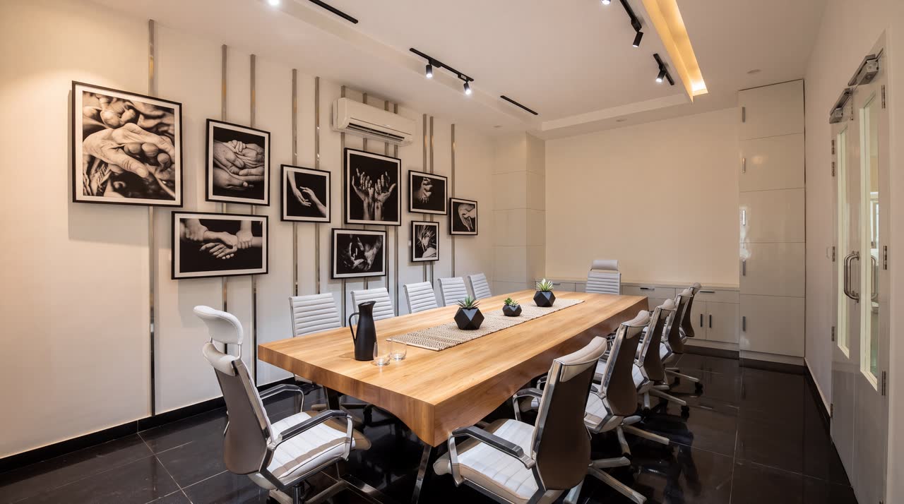 A meeting room with a long live-edge timber table and white mesh chairs, black and white photographs of hands on the left wall, track lighting above and a dark polished floor.